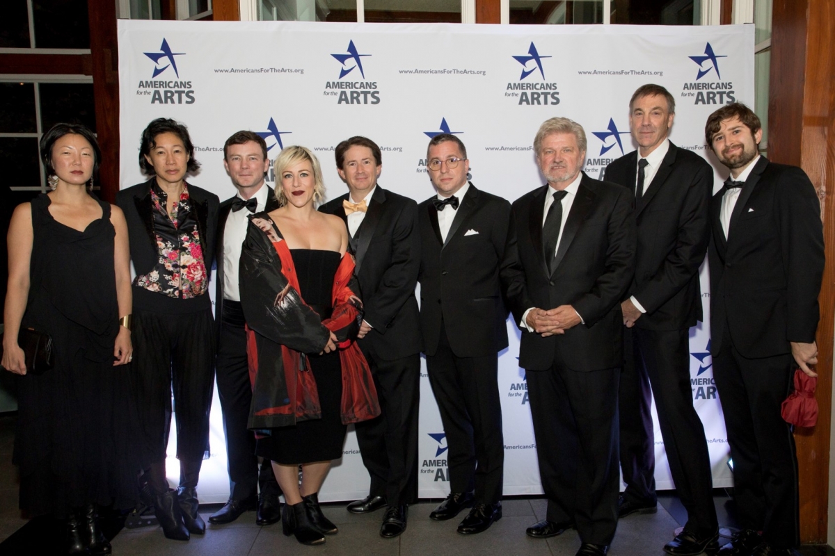 Nine individuals in formal attire pose in front of a step and repeat with the Americans for the Arts logo, featuring a blue star