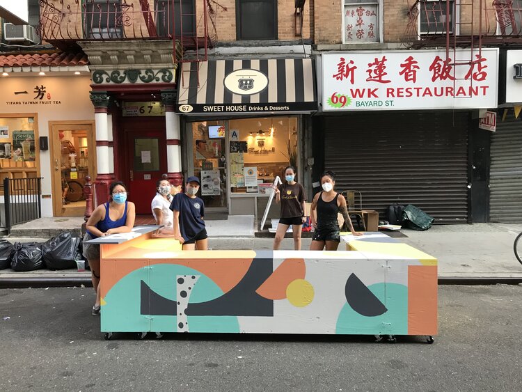 People in masks stand behind a brightly painted dining barrier in front of a restaurant in Chinatown.