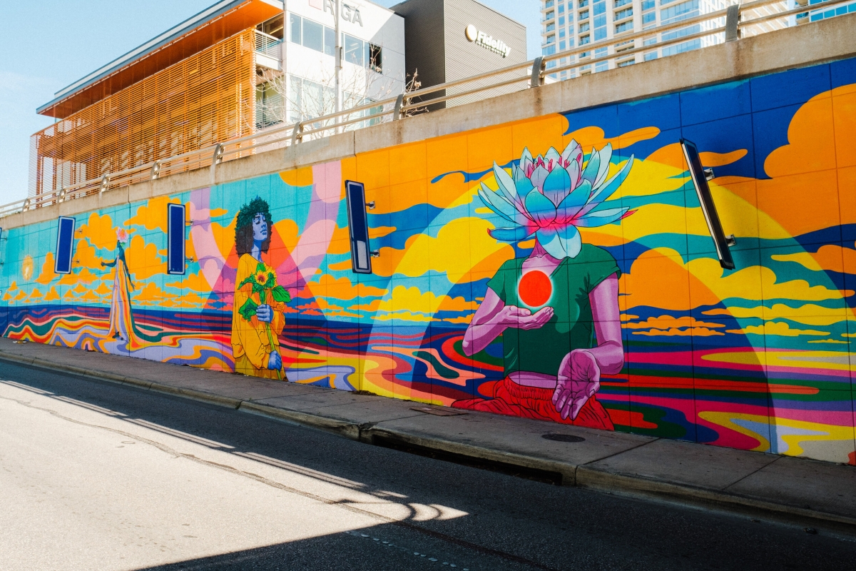 Large outdoor mural in bright sunny colors featuring figures with flowers and faces turned to the sun.