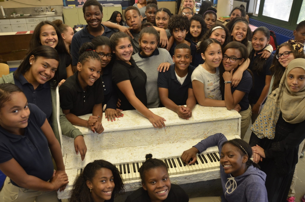 A group of students pose for the camera around a white textured piano.