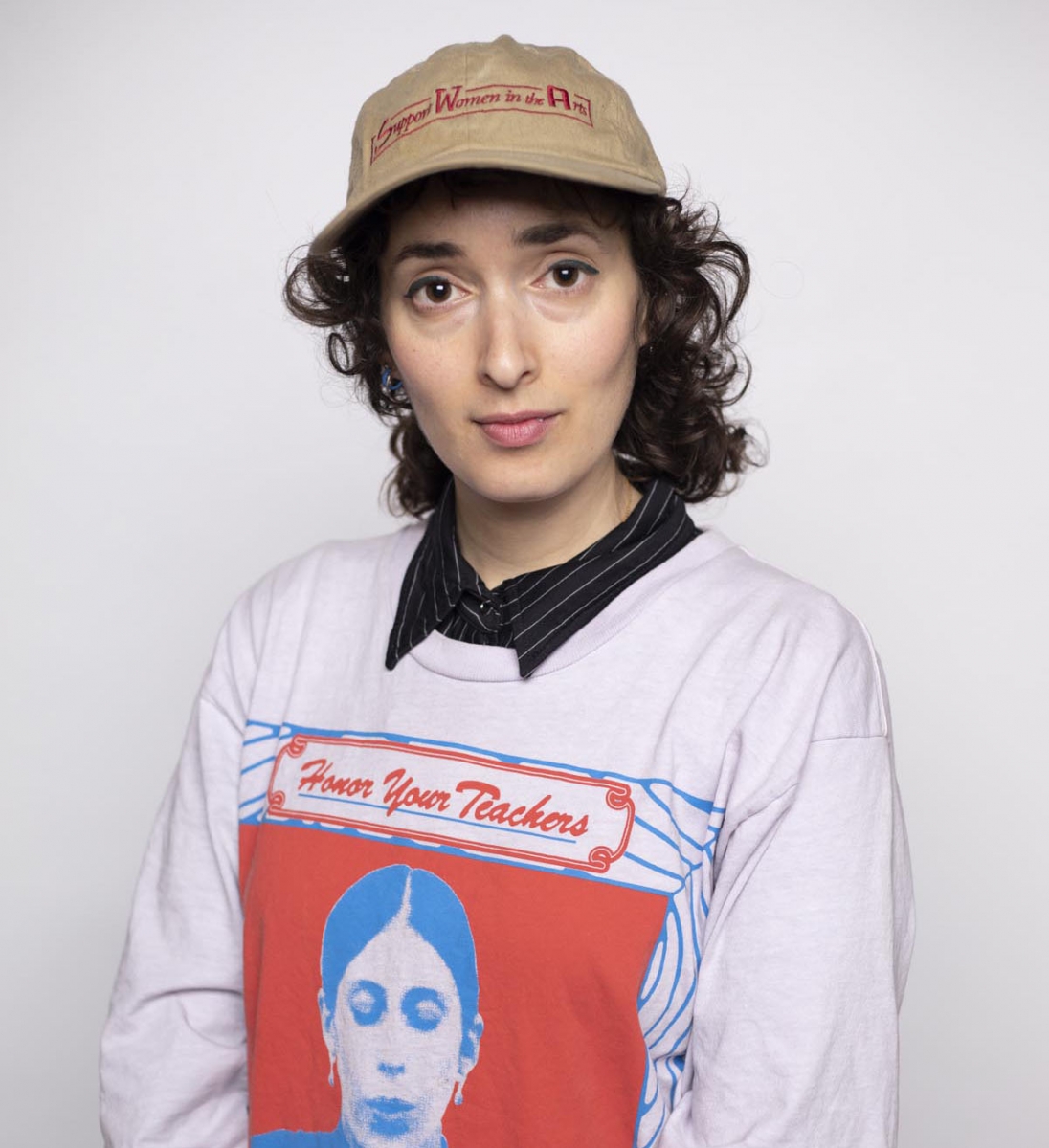 Person with dark curly hair wearing a tan baseball cap and longsleeve white shirt with text Honor Your Teachers and a red and blue silkscreen of a schoolteacher.