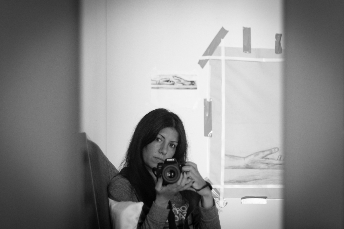 A black and white photo of a person taking their own photo in a mirror. Behind them are drawings of hands on a wall.