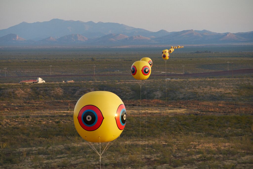 Aerial view of empty land with mountains in the distance. A line of large yellow balloons painted with red, blue, and black eye symbols float high in the sky.