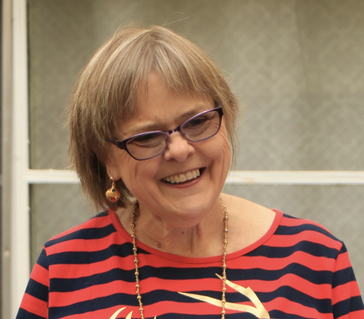 Smiling person with chin-length blonde hair, wearing a red and black horizontally striped shirt, gold necklace and dangling earrings, and purple glasses