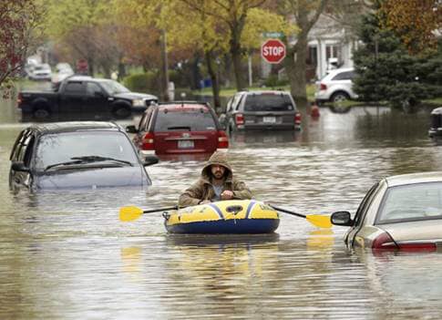 A man wearing a light brown coat with the hood up rows through flooded streets in a blue and yellow rafting tube, while cars, minivans, and a pickup truck try to navigate the street, as water levels reach above the vehicle’s hoods.