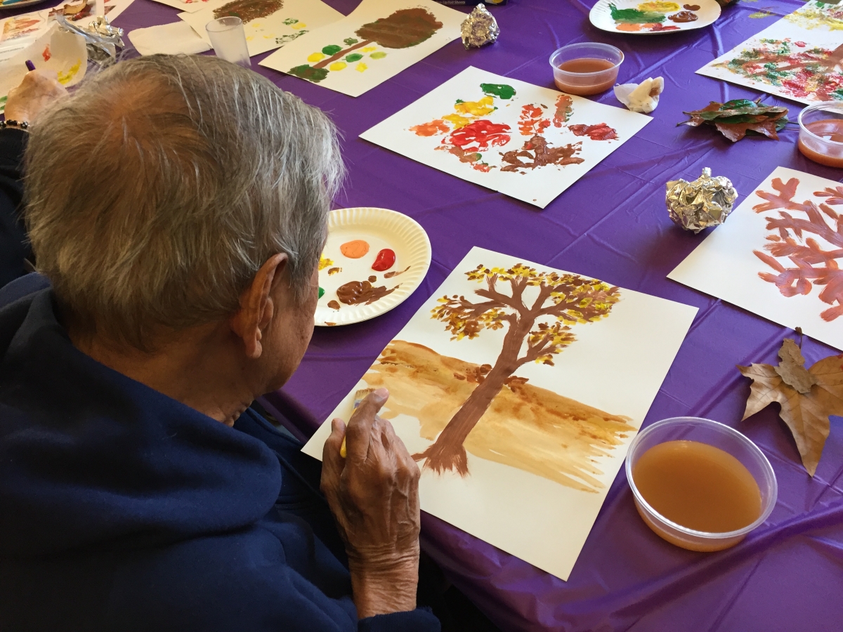 Senior citizen sits at table with purple table cloth and array of watercolor paintings and paints; their piece is of a tree with gold leaves.