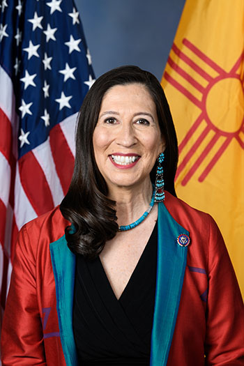 Smiling woman with long dark hair wearing a red silky blazer with turquoise lapels, turquoise necklace, and beaded turquoise dangle earrings, standing in front of the U.S. and New Mexico flags.