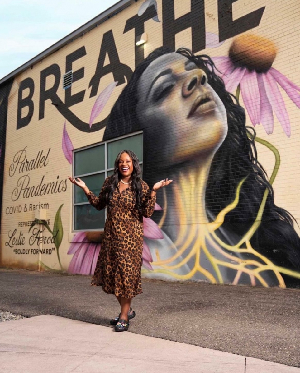 Smiling African American woman with long, wavy dark hair falling below her shoulders, wearing a brown animal-print dress and string of pearls, standing in front of an outdoor mural of a woman with upturned face and closed eyes. The word breathe is in large capital letters and cursive text reads: Parallel Pandemics COVID & Racism, Representative Leslie Herod, Boldly Forward.