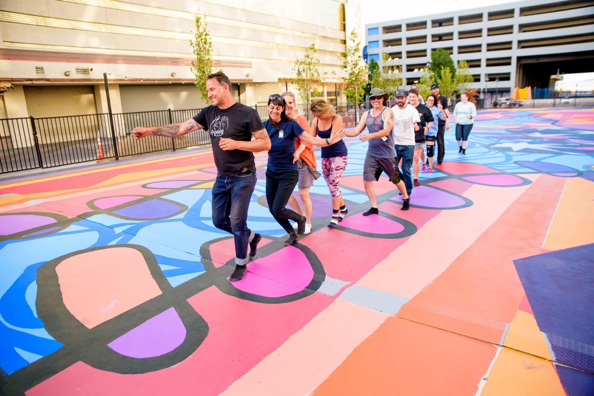 Artist leads volunteers in conga line on outdoor plaza mural brightly painted in shades of pink, blue, peach, orange, purple, and black.