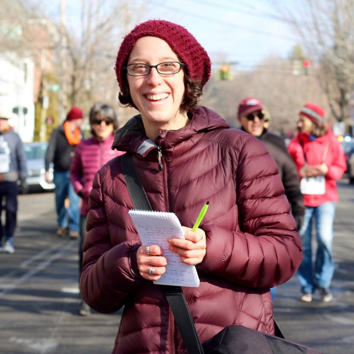 Smiling reporter in the field, wearing dark-rimmed glasses, red knit hat and puffy maroon coat, with black bag slung over shoulder, and holding notepad and pen.