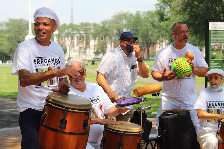 Drummers play congas, claves, shekere, and guiro, dressed in white Bregamos Community Theater t-shirts, perform outdoors in the park on a sunny day. 