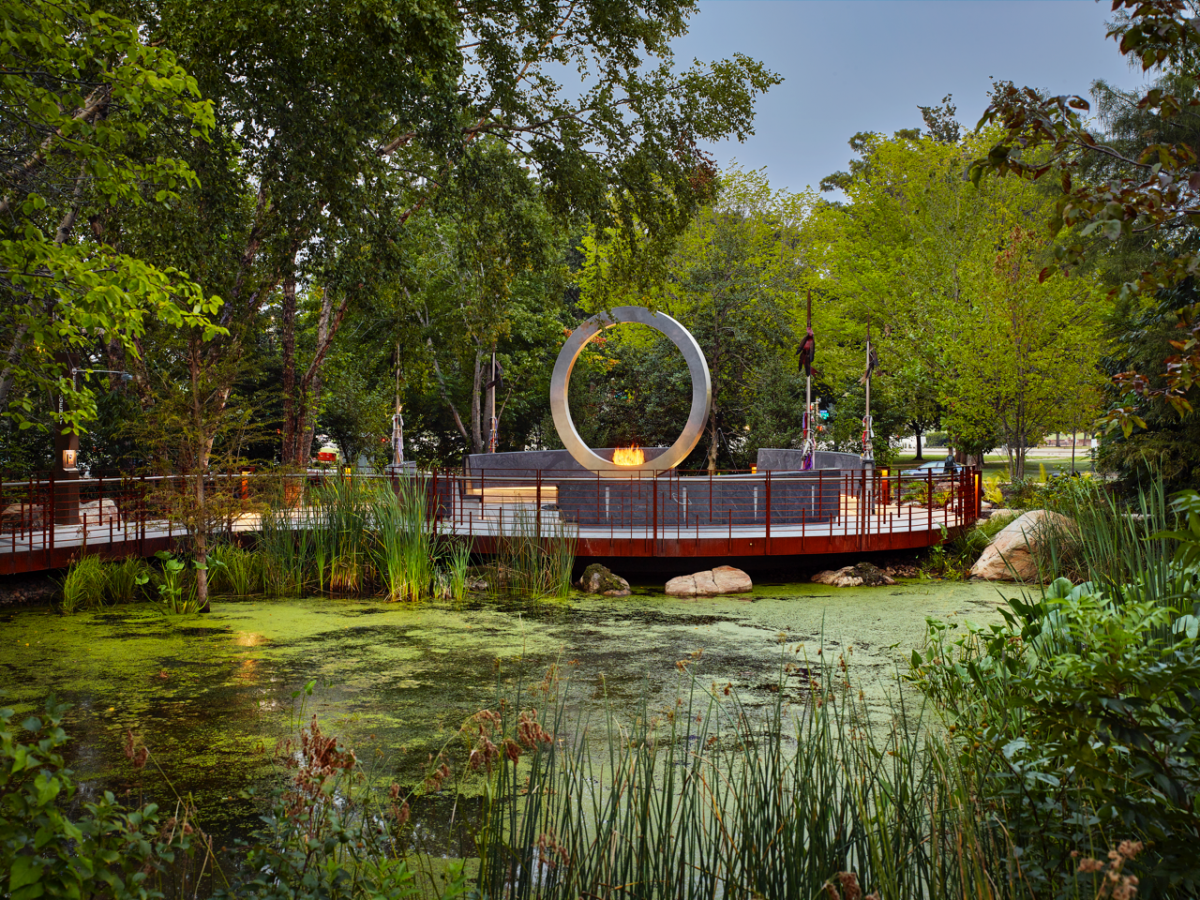 A medium distance view of the memorial. A large upright metal ring with flames in the center rises from the center of a platform surrounded by a marshy pond. The entire memorial is surrounded by trees.