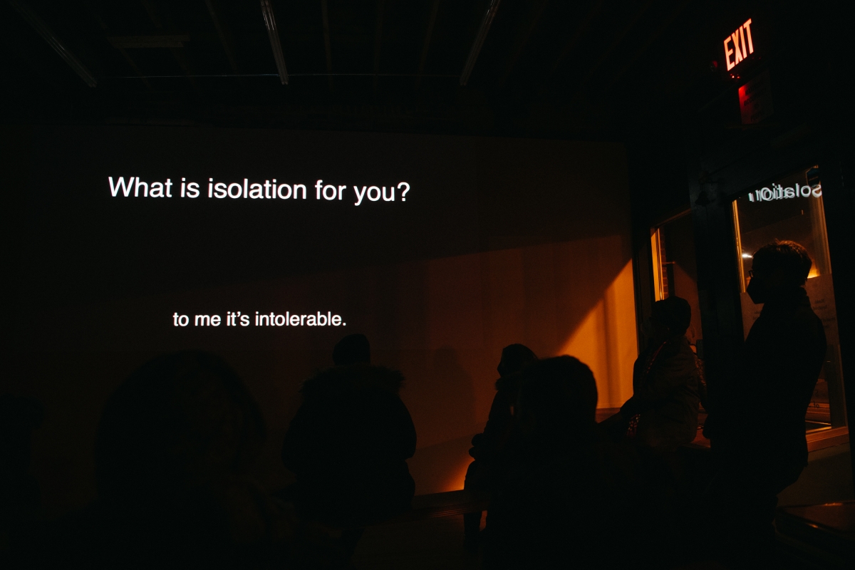 A dark gallery room at nighttime with gallery attendees viewing an open text caption video, with white text against a black background. The text reads “What is isolation for you?”, “to me it's intolerable.”