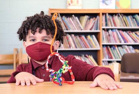 A student with curly hair wearing a red mask and shirt sits at a wood table with a sculpture made of pipe cleaners.