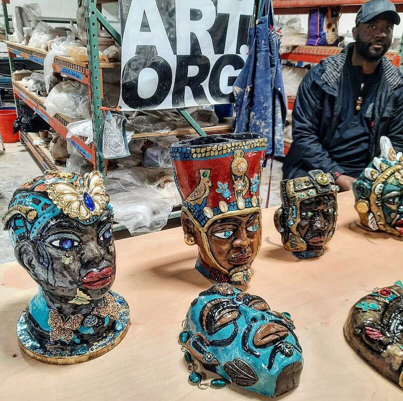 A person sits behind a table on which sit medium sized ceramic sculptures of heads in the style of African royalty and decorative masks.