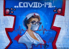 Graffiti style mural painted on a brick wall showing a nurse in traditional white uniform and a white mask with a heartbeat graph behind them and COVID-19 above them. 